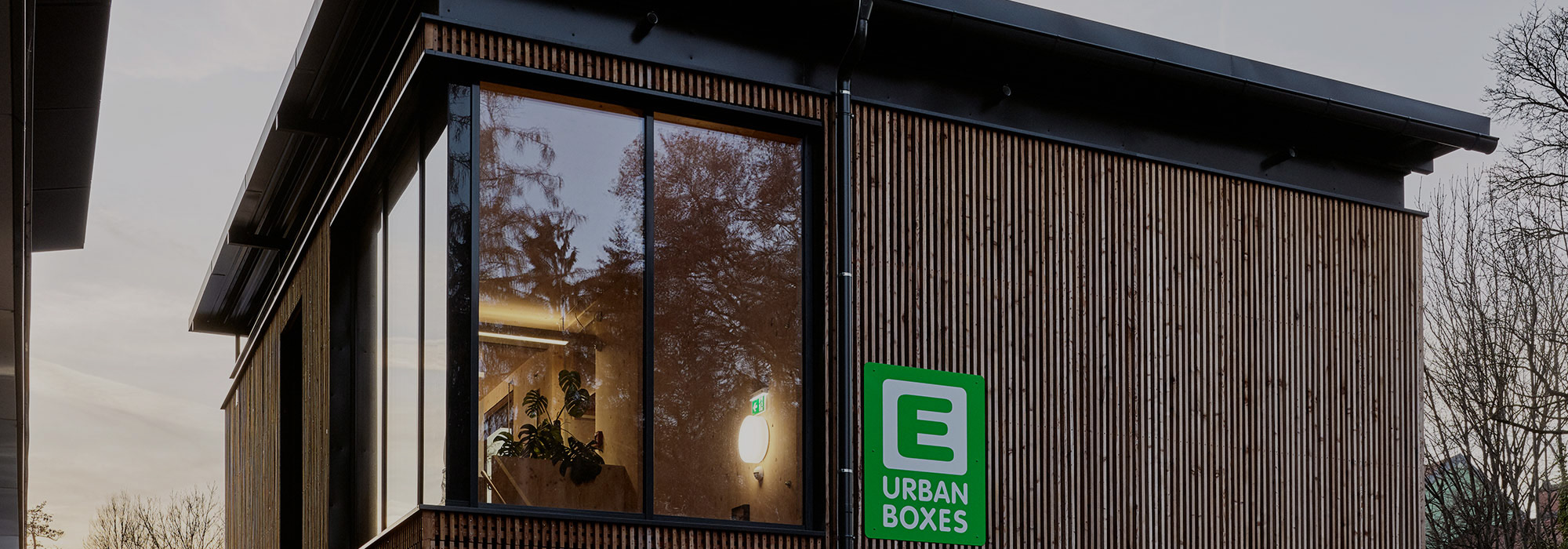 Ein modernes Holzhaus, eine sogenannte Urban Box, ist in der Abenddämmerung zu sehen.